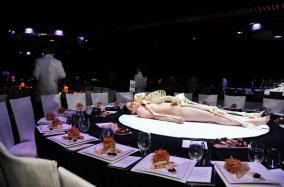 Performance actors in training lying naked under plastic skeletons revolving on the $100,000 dinner tables. 